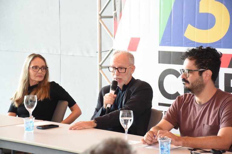 Foto de três pessoas sentadas à mesa. Da esquerda para direita: a produtora Cláudia da Natividade, mulher de pele clara e cabelos longos loiros, usando óculos. Ela observa o diretor Marcos Jorge, homem de pele clara e cabelos e barba grisalhos sentado ao centro. Ele usa óculos e segura um microfone. Ele fala ao público. À direita, o curador do festival Edu Fernandes, homem de pele clara e cabelos e barba preta. Ele usa óculos. Ao fundo, um banner com a identidade visual da 30ª edição do CINE/PE.