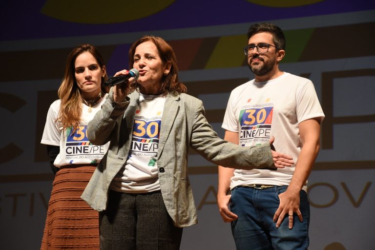 Foto. Da esquerda para direita: Patricia, Sandra e Vitor Bertini no palco. Todos usam a camisa da 30ª do Cine/PE