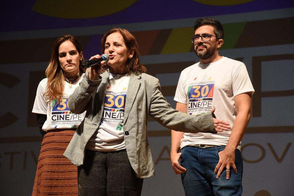 Foto. Da esquerda para direita: Patricia, Sandra e Vitor Bertini no palco. Todos usam a camisa da 30ª do Cine/PE
