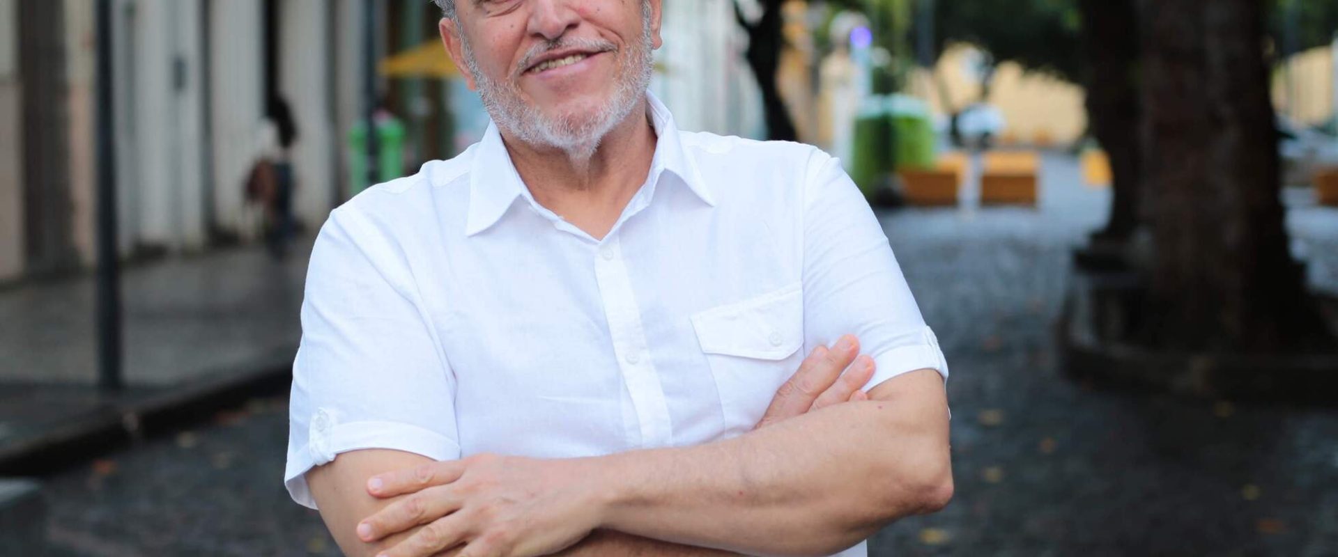 Foto de Alfredo Bertini, homem de pele clara de cabelos e barbas grisalhos. Ele está com os braços cruzados e veste uma camisa brancas de manga média. Ao fundo, rua arborizada.