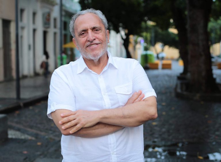 Foto de Alfredo Bertini, homem de pele clara de cabelos e barbas grisalhos. Ele está com os braços cruzados e veste uma camisa brancas de manga média. Ao fundo, rua arborizada.